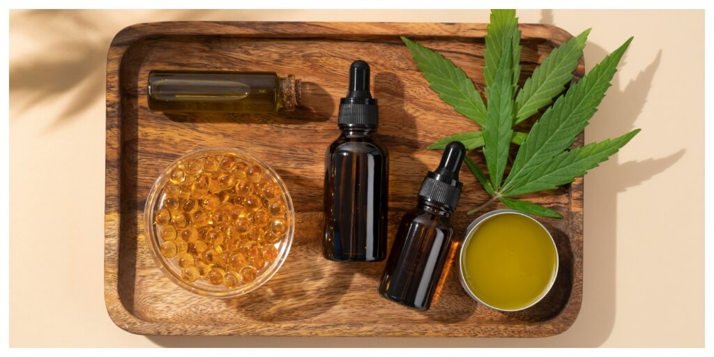 CBD oil products on a wooden tray with hemp leaf – capsules, tinctures, balm and glass bottle