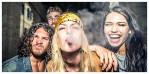 four friends celebrating outside, blonde girl with a bandana is smoking, girl on her right is laughing. Guy on the left is making a weird face and a guy in the back is smiling.