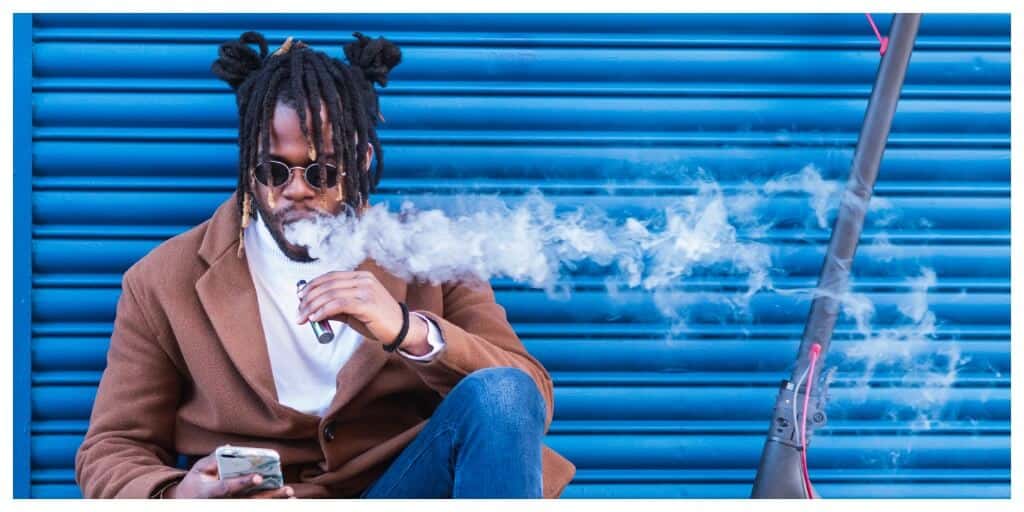Hipster black man with electronic cigarette on his phone in front of a blue garage door next to an electronic scooter