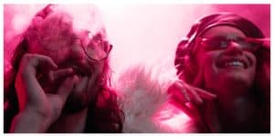 panoramic shot of man smoking marijuana joint near smiling girl in nightclub under red lights