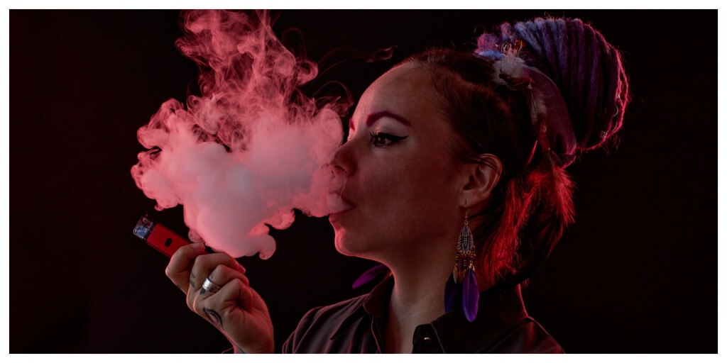 Young woman with feather earings blowing smoke out of her mouth whilst holding a vape in front of a dark background with red neon shades